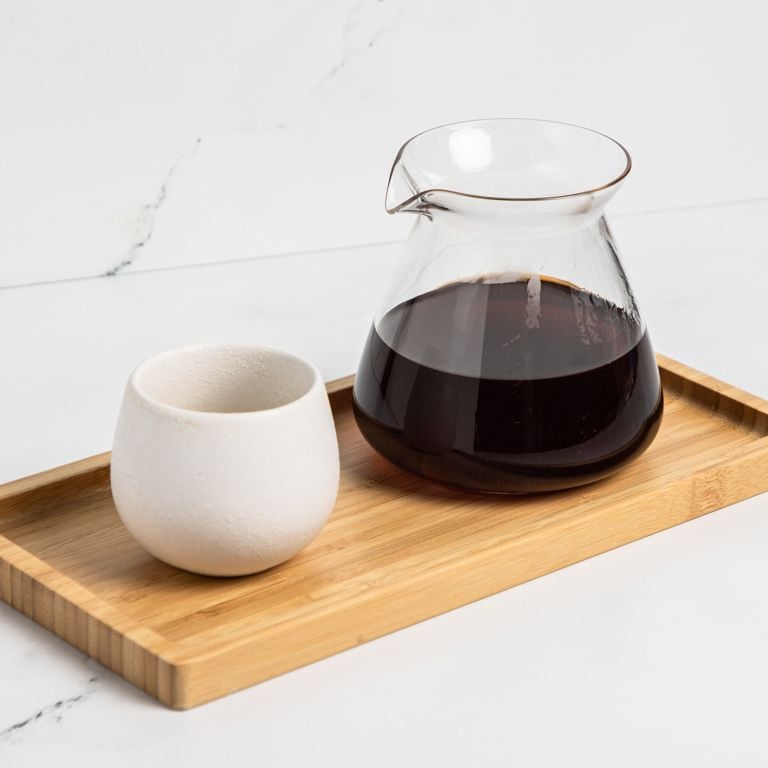 Menu – Pour Over – Board A carafe of pour over coffee next to a cup. Both sit on a wooden board in front of a marbled white background.
